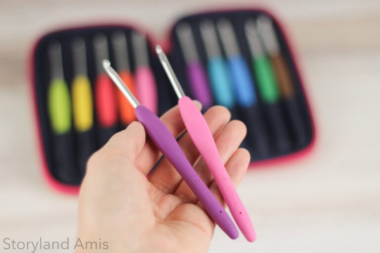 Hand holding up two crochet hooks. One is purple and the other is pink.