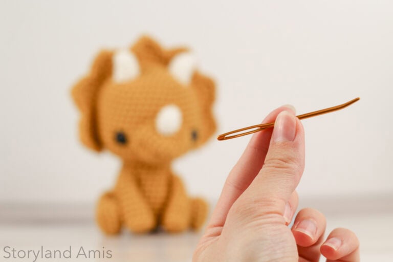 Hand holding up a bent tip tapestry needle. A yellow crocheted triceratops is in the background.