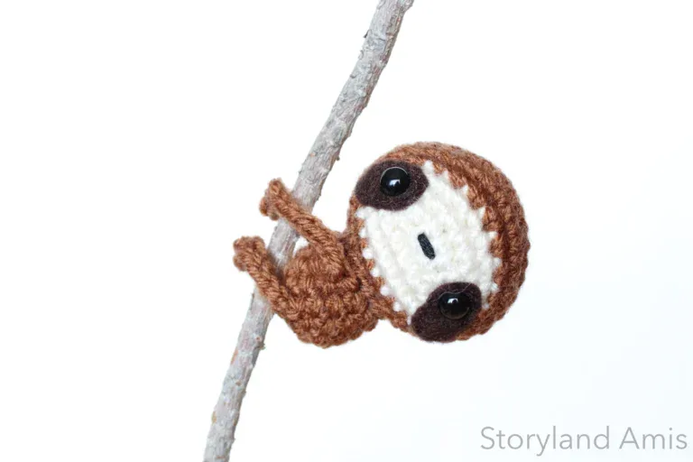 A brown crocheted sloth hangs from a small tree branch against a white background.