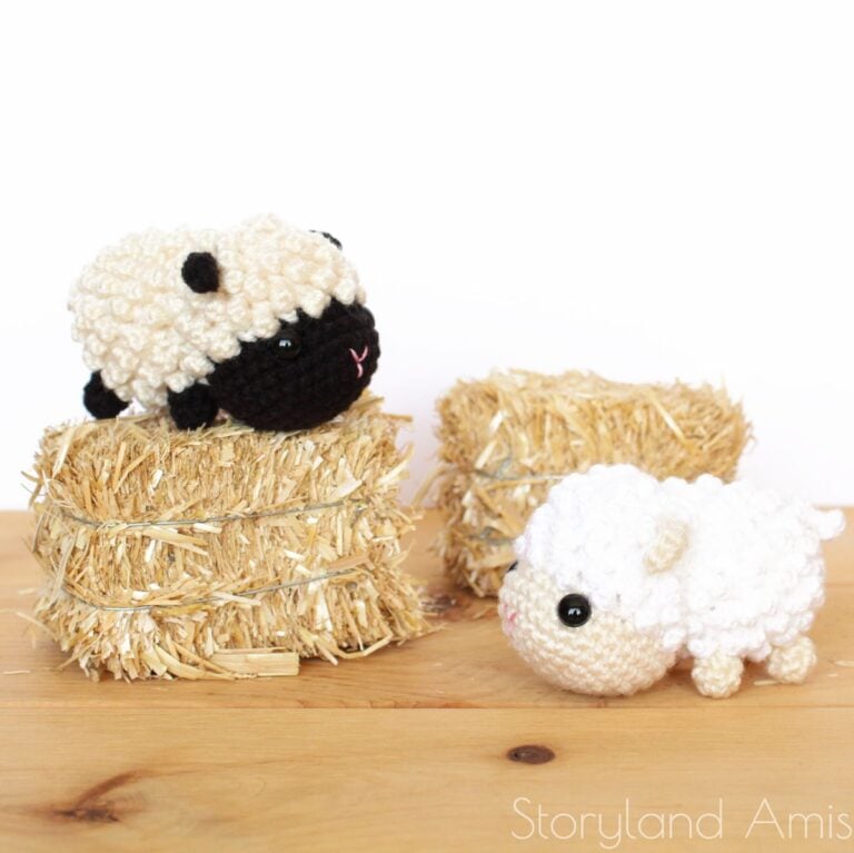 A black crocheted lamb amigurumi sits atop a hay bale while a second crocheted white lamb stands in front of a hay bale.