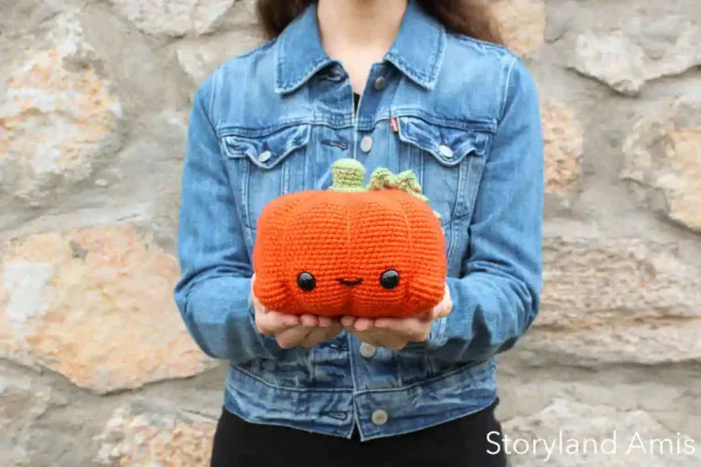 A girl stands against a stone wall holding a crocheted pumpkin.