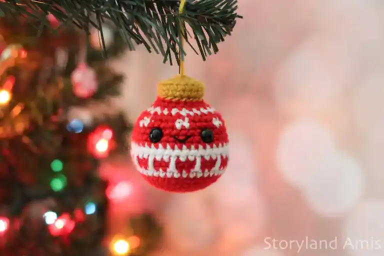 A crocheted Christmas ornament hangs from the a branch with twinkling lights in the background.