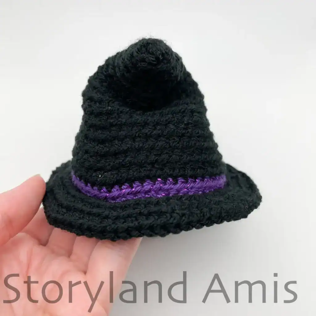 A hand holds a black, crocheted witch hat for an amigurumi ghost. The hat is shown from the side with the top of the hat bent to the side.