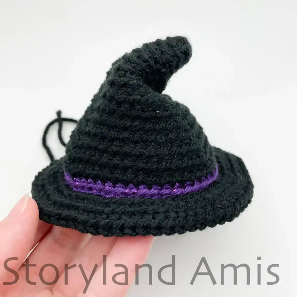 A hand holds a black, crocheted witch hat for an amigurumi ghost. The hat is shown from the front with the top of the hat bent to the side.