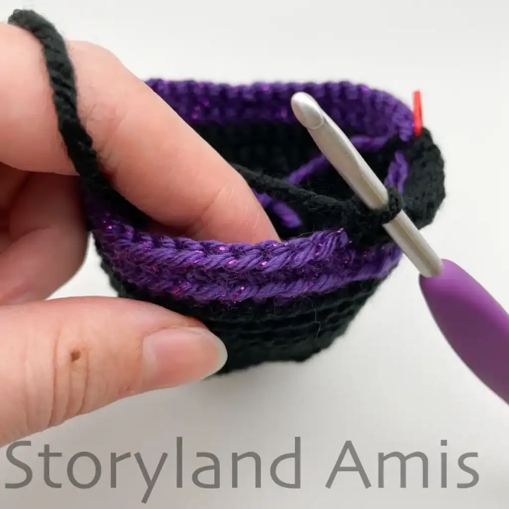Close-up of crochet ghost hat in progress, showing the making of the hat brim. A hand holds the hat steady, and a purple crochet hook is inserted into the first stitch.