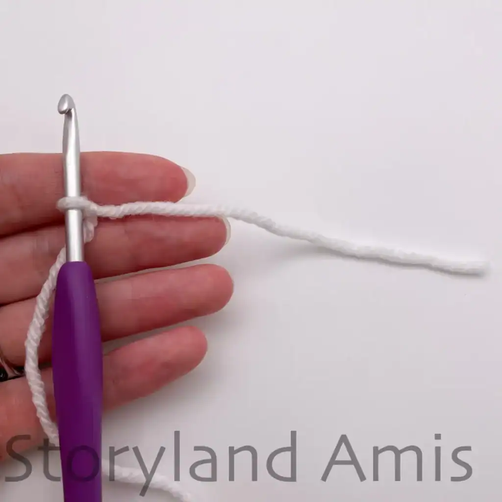 A hand holds a purple crochet hook with white yarn looped around it.