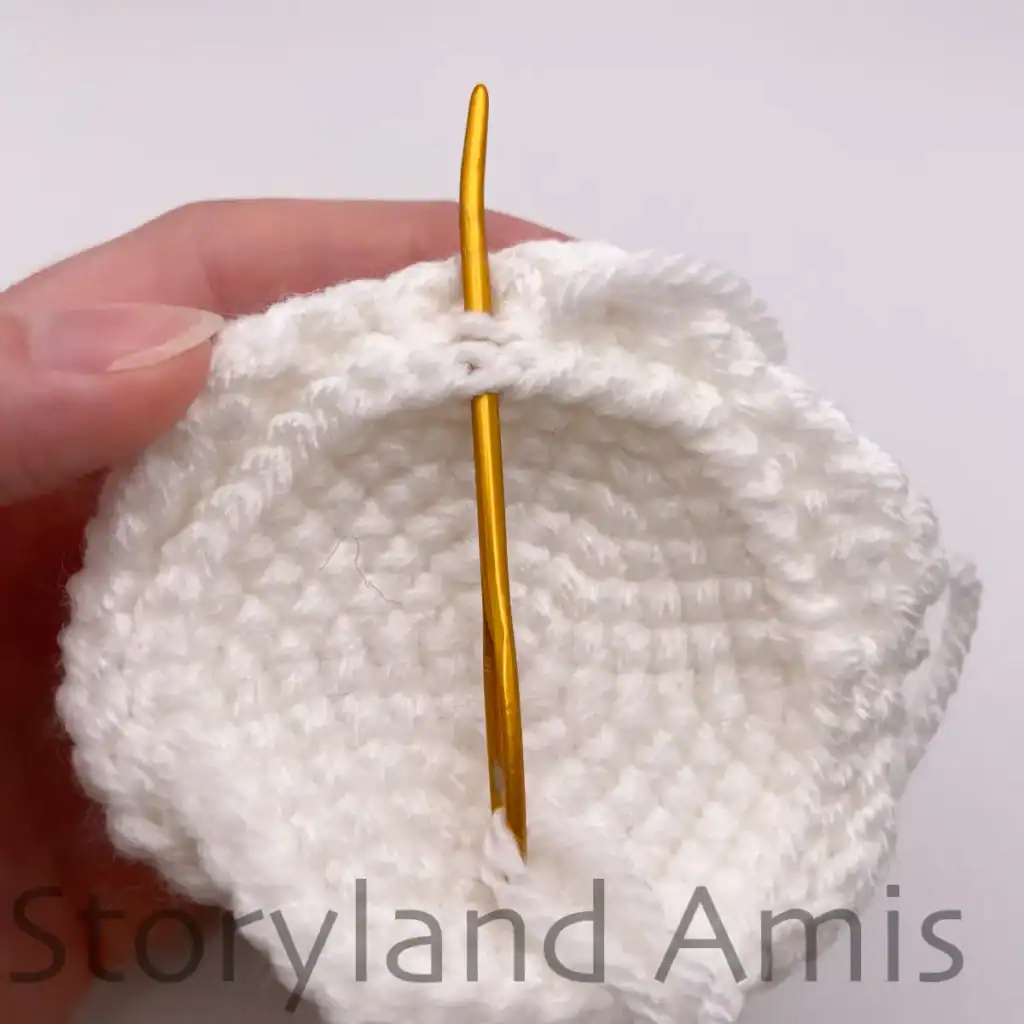 A hand demonstrates how to sew a crocheted bottom piece to the body of an amigurumi ghost using a tapestry needle.