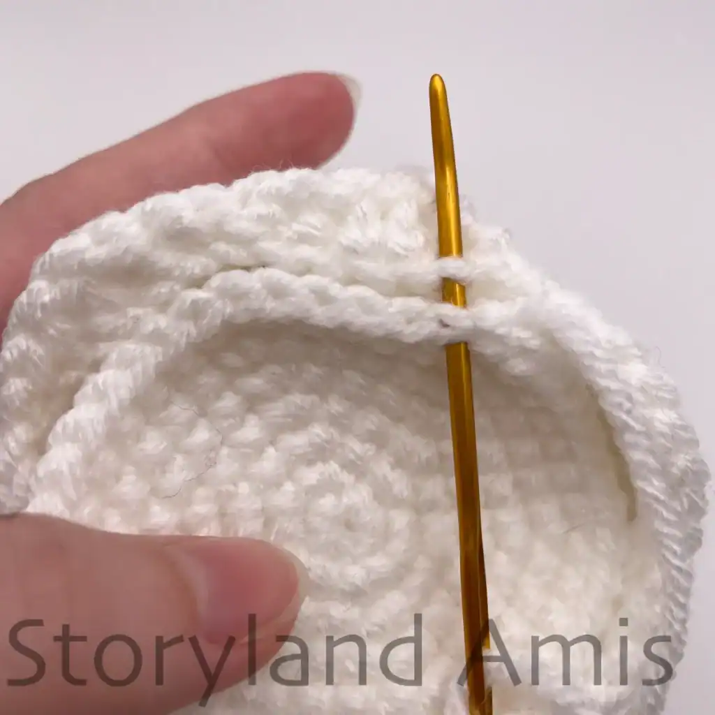 A hand demonstrates how to sew a crocheted bottom piece to the body of an amigurumi ghost using a tapestry needle.