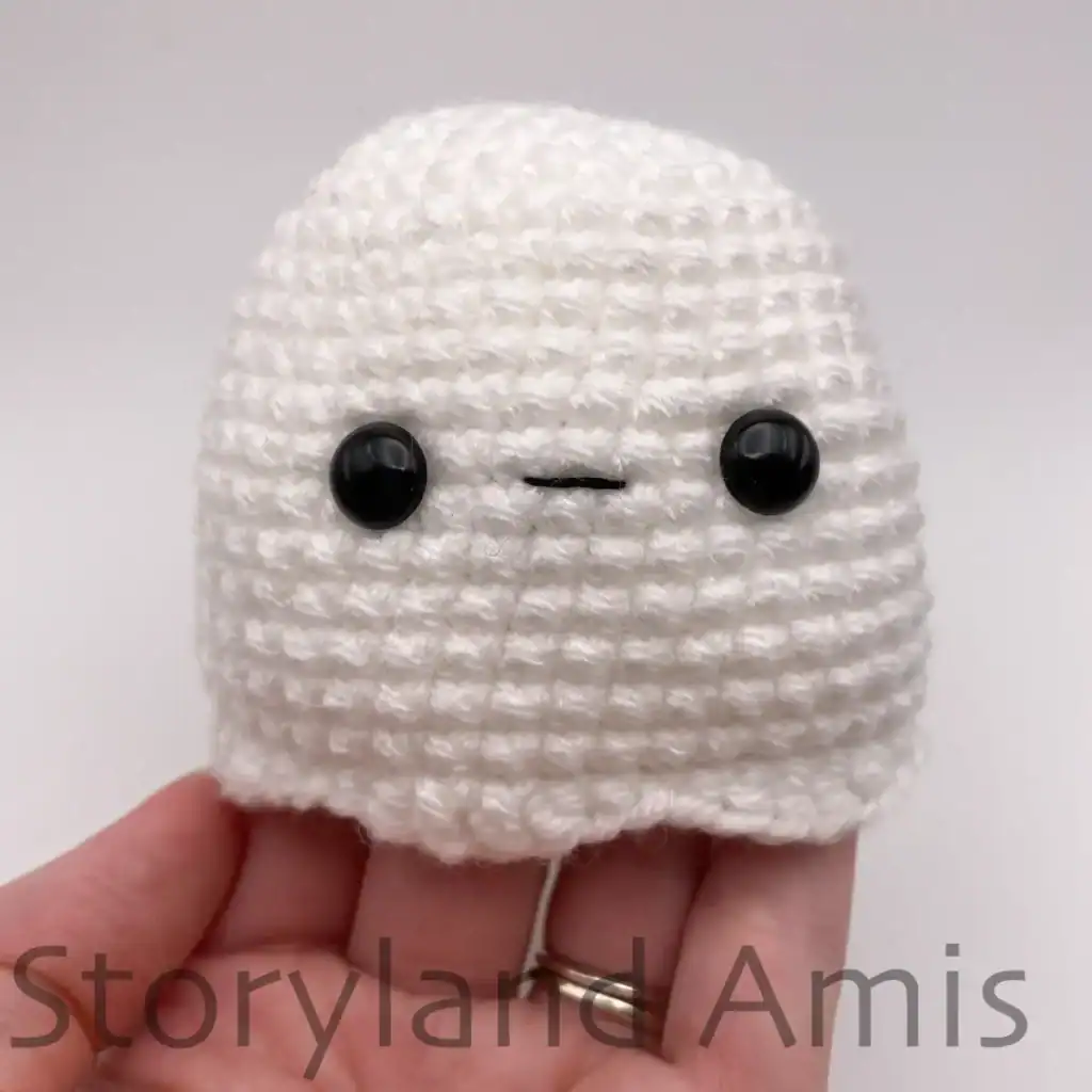 A hand holds up a partially finished amigurumi ghost showing where to embroider the mouth between the eyes.