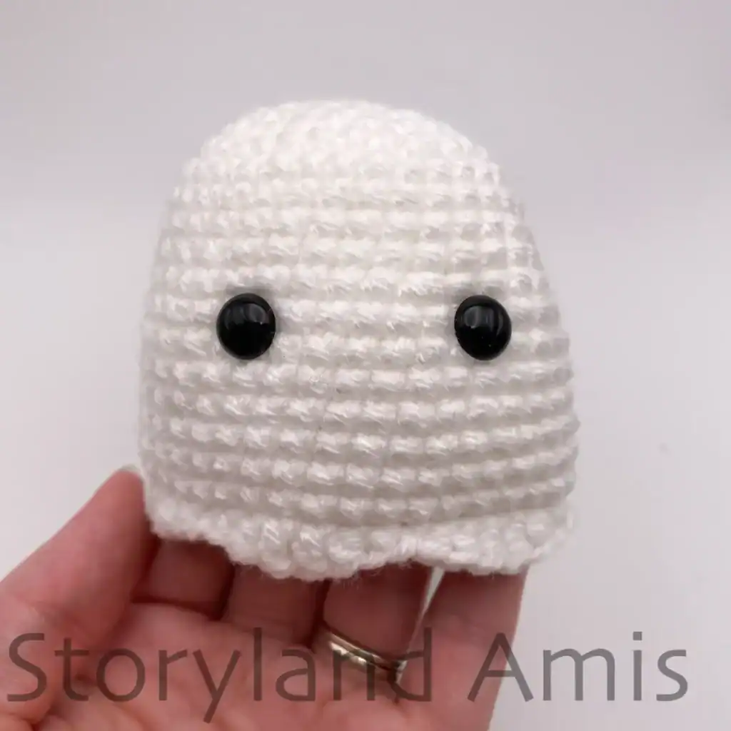 A hand holds up a partially finished amigurumi ghost showing where to position the safety eyes on the face.