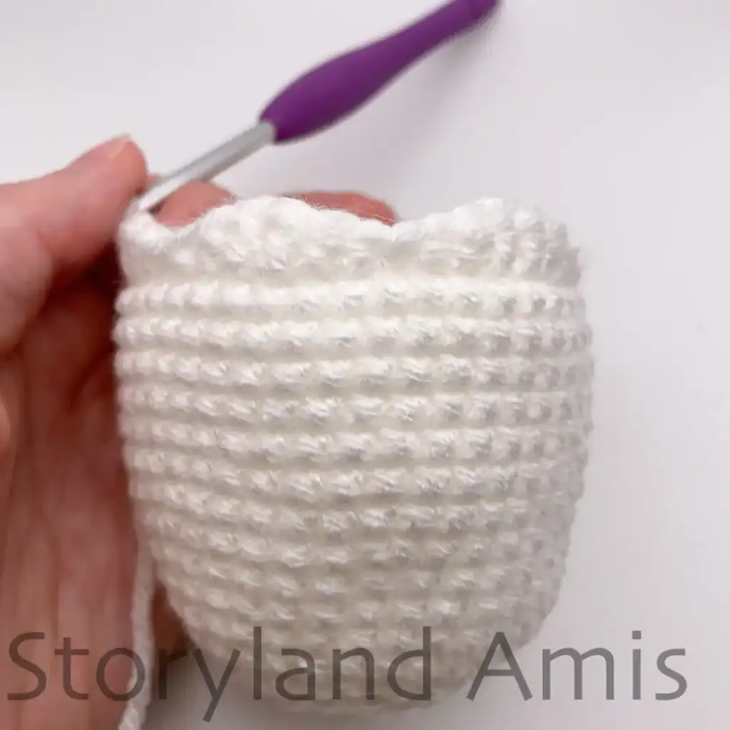 Close-up of a crochet ghost body in progress, showing the scalloped fringe formed at the bottom edge using various stitches. A crochet hook with purple handle is inserted into a stitch at the top, with a hand holding the work steady.
