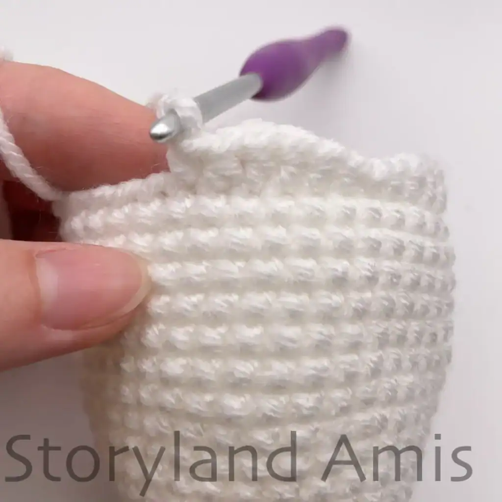 Close-up of a crochet ghost body in progress, showing the scalloped fringe formed at the bottom edge using various stitches. A crochet hook with purple handle is inserted into a stitch at the top, with a finger holding the work steady.