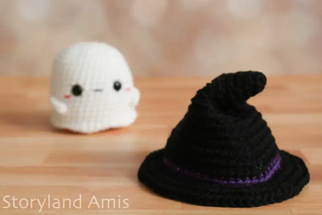 A crocheted black witch hat sits on a wooden table. An amigurumi ghost sits in the background.