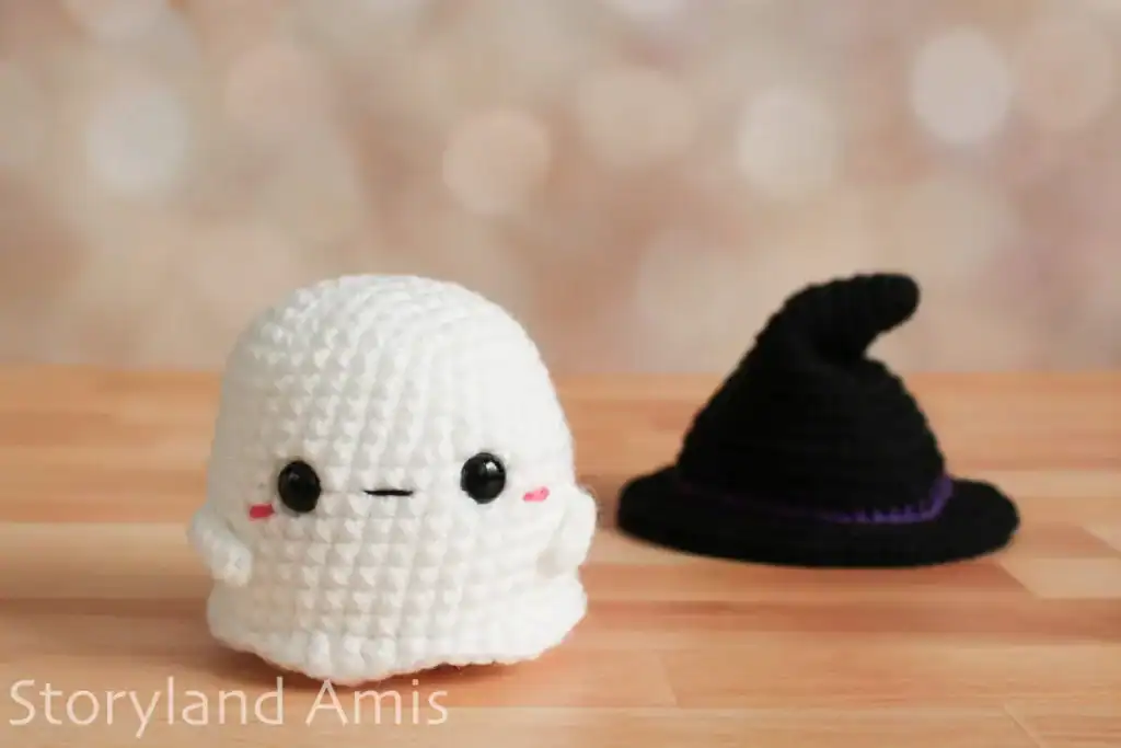 A crocheted baby ghost sits on a wooden table with a black crocheted witch hat in the background.