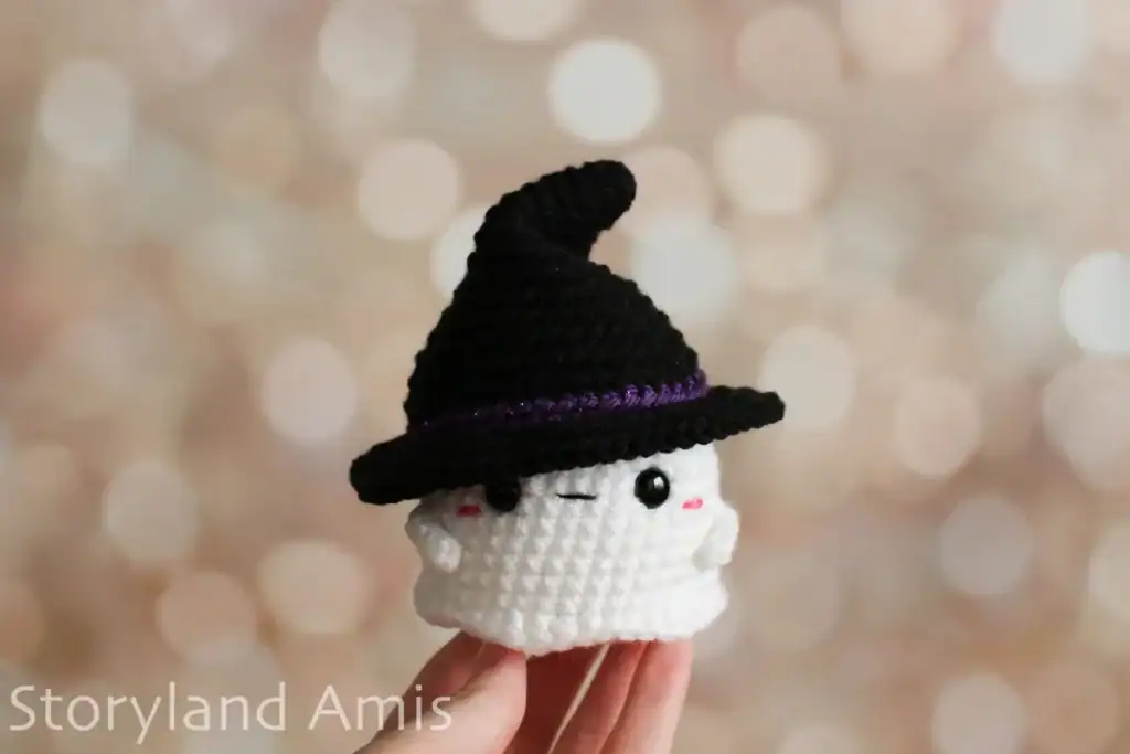 A hand holds up a mini amigurumi ghost wearing a black witch hat.