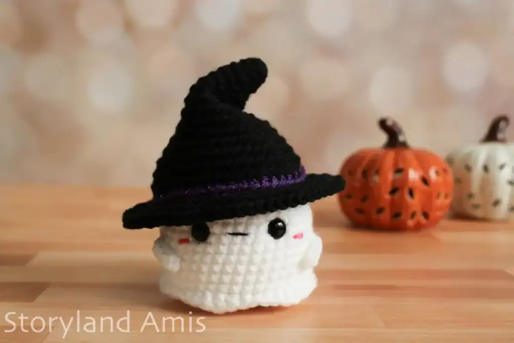 A crocheted baby ghost amigurumi wearing a witch hat sits on a wooden table with two ceramic pumpkins behind it.