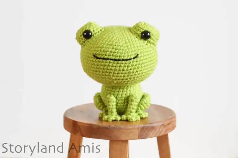 A green amigurumi frog sits on a small wooden stool.