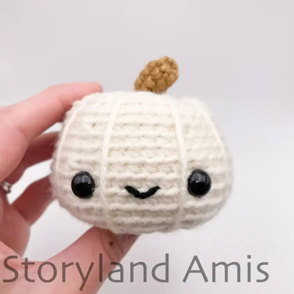 A hand holds a mini white amigurumi pumpkin.
