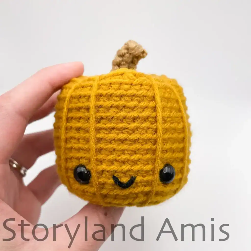 A hand holds a mini yellow amigurumi pumpkin.