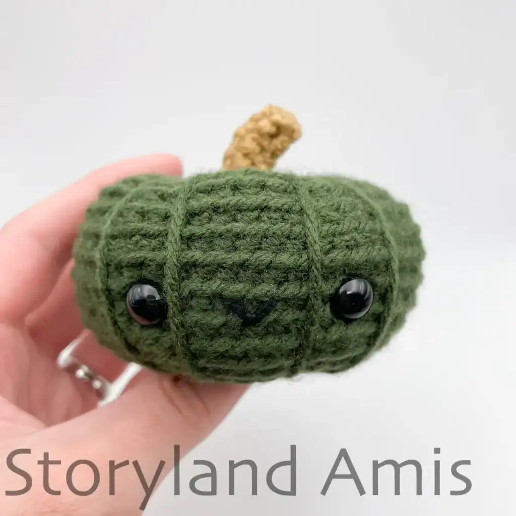 A hand holds a mini green amigurumi pumpkin.
