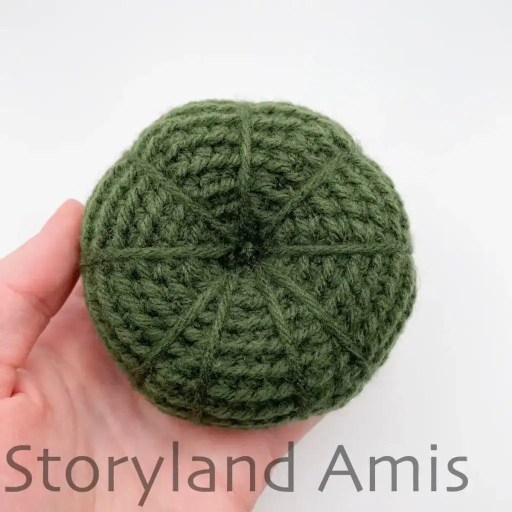 A hand holds a partially finished green amigurumi pumpkin shown from the top.