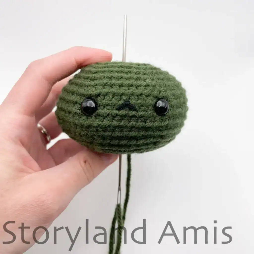 A hand holds a partially finished amigurumi pumpkin upside down, demonstrating how to pull the tapestry needle through the center of the body.