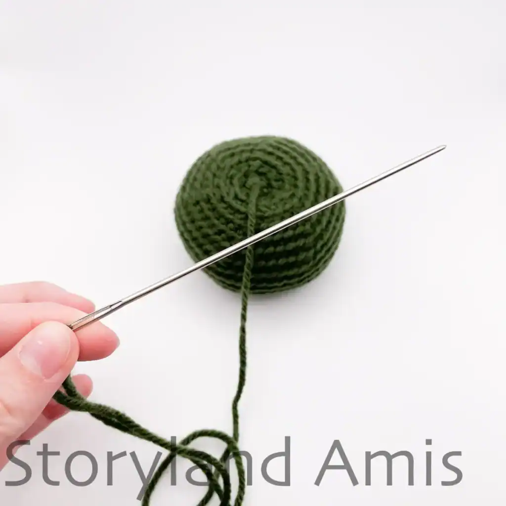 A hand holds an extra-long tapestry sewing needle threaded with green yarn. The green yarn is attached to a partially finished amigurumi pumpkin.