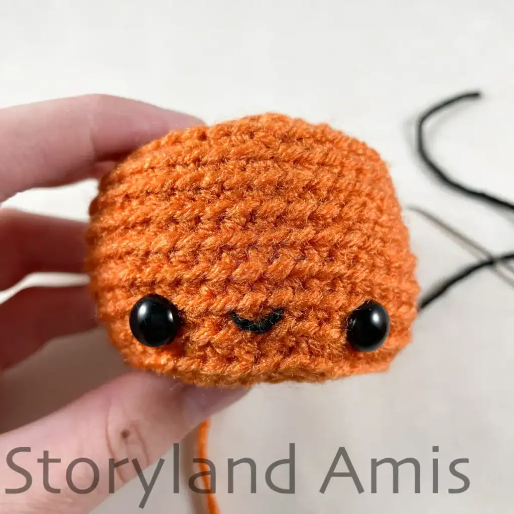 A hand holds up a partially finished amigurumi pumpkin, showing where mouth should be embroidered between the eyes.