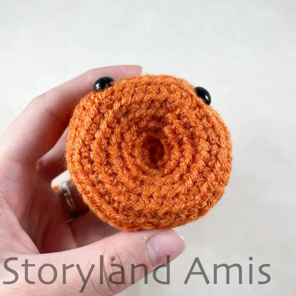 A hand holds up an orange crocheted pumpkin, shown from the bottom.