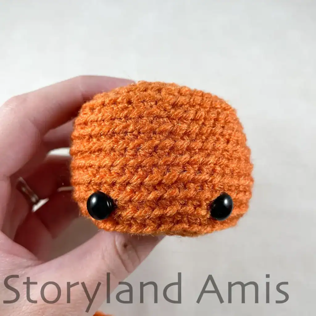 A hand holds up a partially finished amigurumi pumpkin, showing where the safety eyes are to be placed on the face.