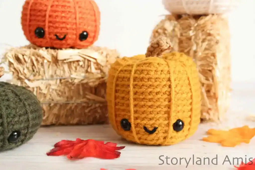 Close-up of cute crocheted pumpkins in fall colors with smiling faces, surrounded by straw bales and autumn leaves.