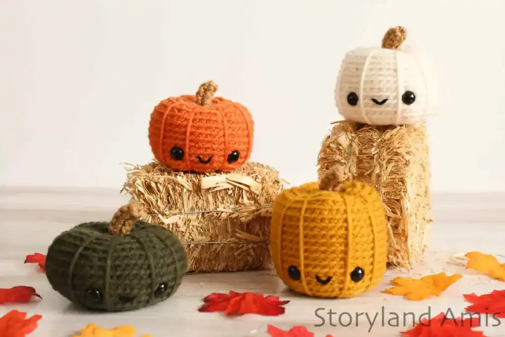 Four colorful amigurumi pumpkins with smiling faces, crocheted in orange, white, mustard yellow, and dark green yarn, are displayed on small hay bales and surrounded by decorative autumn leaves.