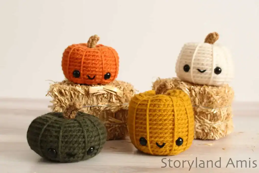 Four colorful amigurumi pumpkins with smiling faces, crocheted in orange, white, mustard yellow, and dark green yarn, are displayed on small hay bales.