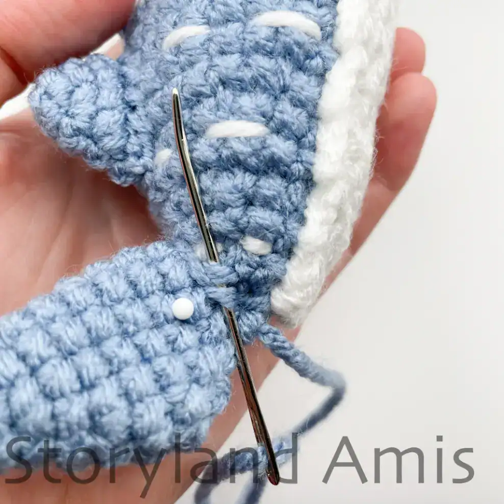 A hand demonstrates how to sew a tail fin onto an amigurumi whale shark.