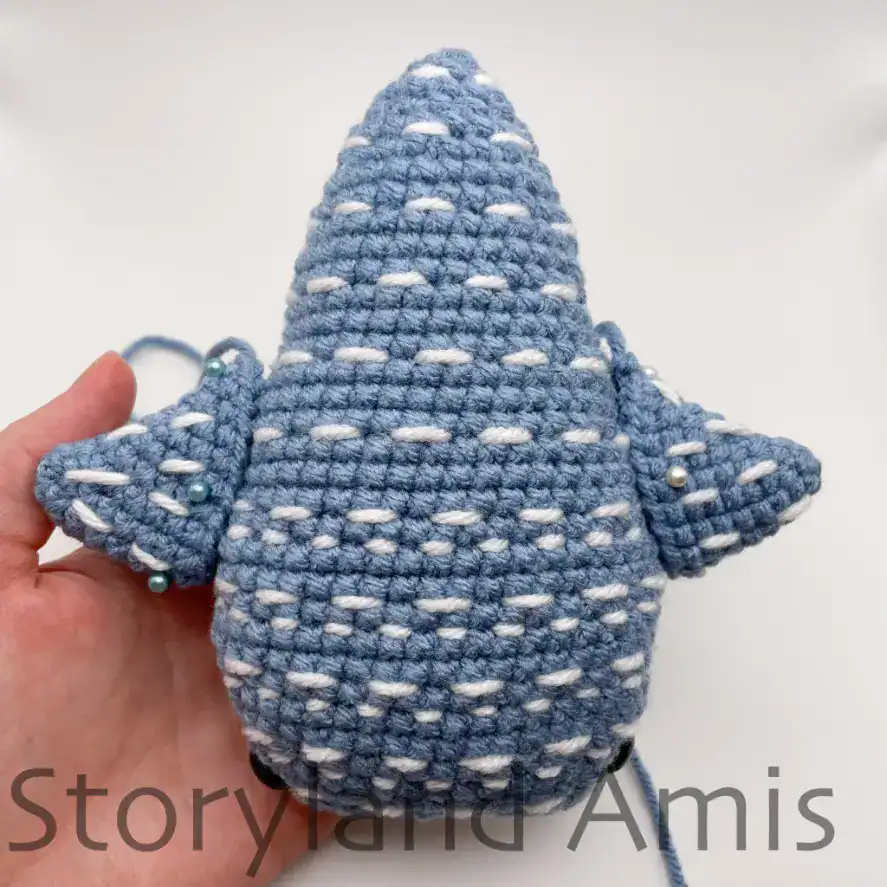 A hand holds a partially finished amigurumi whale shark, showing where to place the fins.