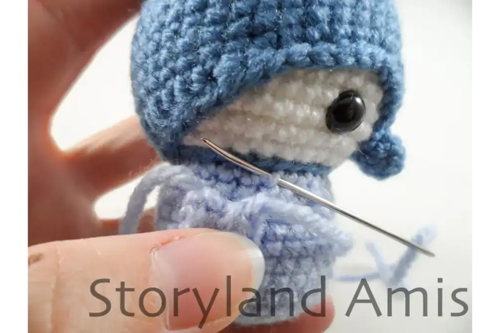 A close-up of a hand demonstrating how to sew a wing to the body of an amigurumi penguin using a tapestry needle.
