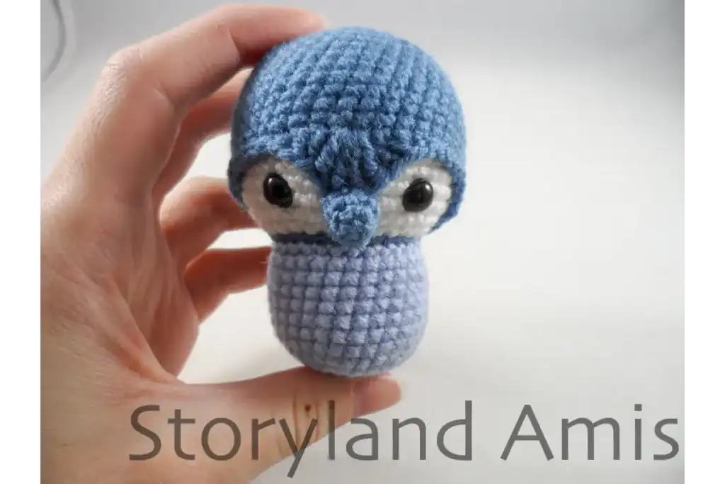A hand holds up a partially finished blue amigurumi baby penguin.