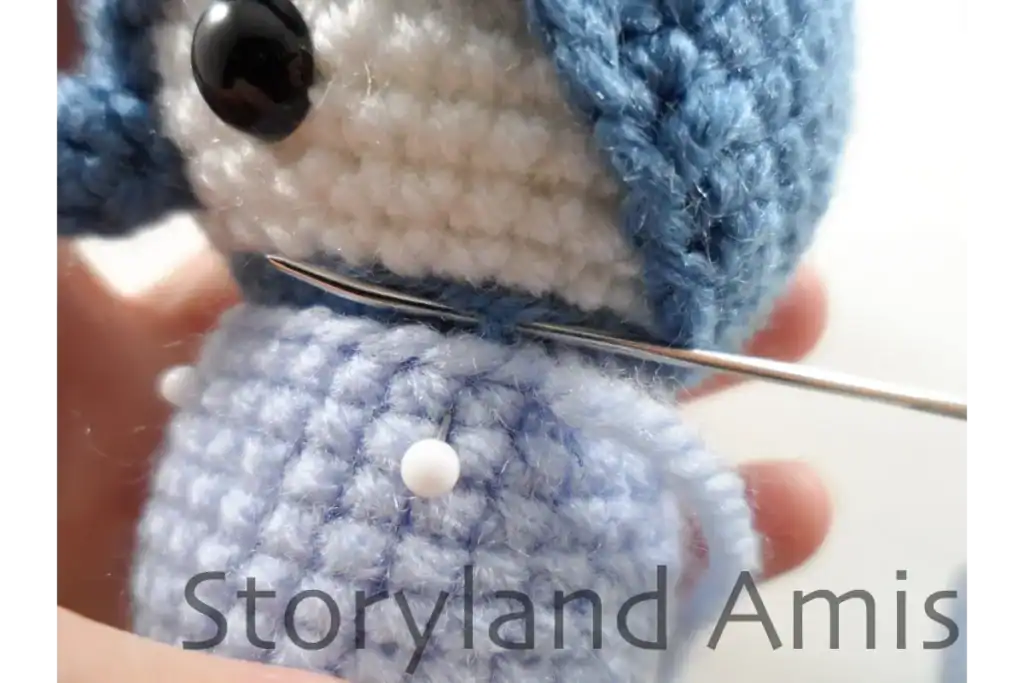 A close-up of a blue, amigurumi penguin showing the body being sewn to the head using a tapestry needle.