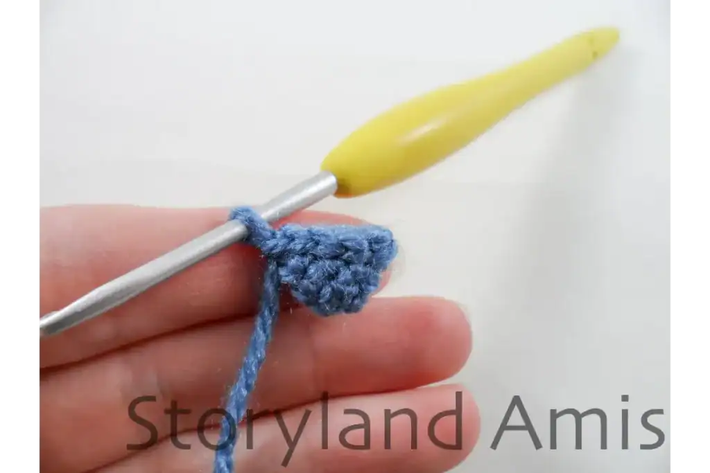 A hand holds a small, blue crocheted penguin foot with a yellow crochet hook inserted into a stitch in the corner of the foot.