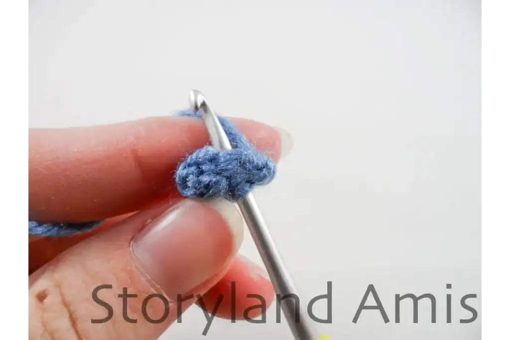 A hand holds a small, blue crocheted penguin foot. A crochet hook is inserted into the stitches of the foot, showing how to crochet the foot closed.