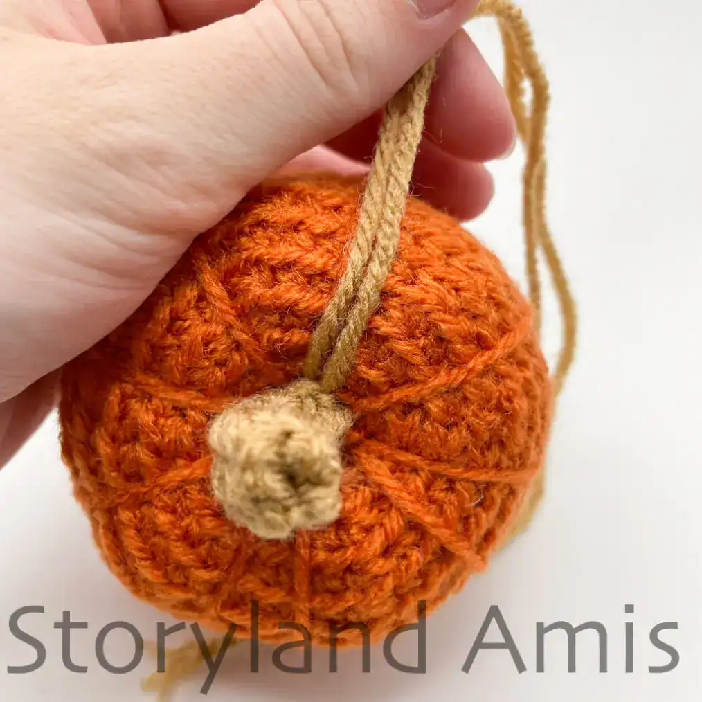 A close-up of the top of an amigurumi pumpkin with a brown stem. A hand holds the two yarn tails still attached to the stem that were used to sew the stem to the pumpkin.