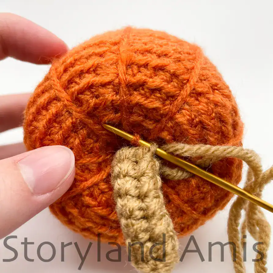 Close-up of an orange crocheted pumpkin with a brown stem being sewn onto the top using a gold yarn needle and matching brown yarn.