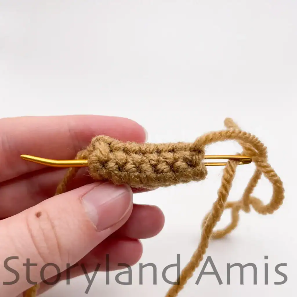 Close-up of a hand guiding a gold yarn needle through the center of a crocheted brown stem piece, passing from top to bottom as part of the assembly for an amigurumi pumpkin.
