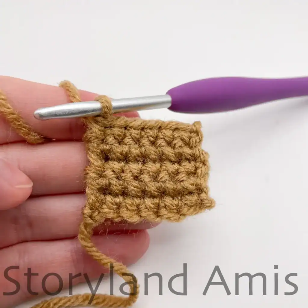 A hand holds a crocheted work-in-progress pumpkin stem. A purple crochet hook is inserted in the top corner of the pumpkin stem.