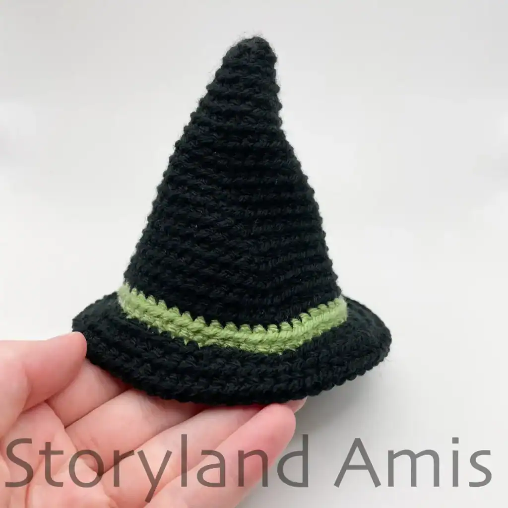 A hand holds a completed crocheted witch's hat. The hat is black with a green band around the brim.