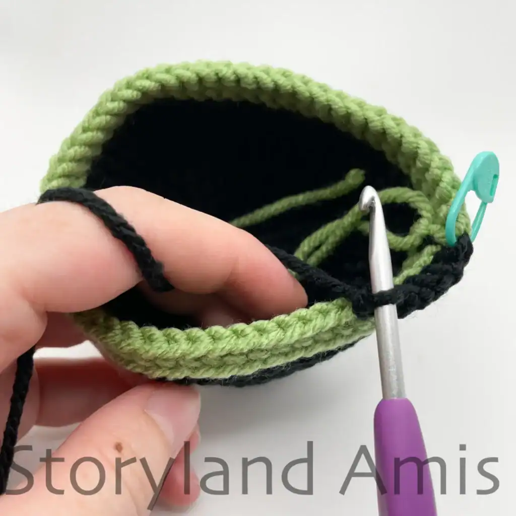 A close-up of the edge of a crocheted hat shows the brim of the hat being made. A purple crochet hook is inserted in a stitch of the hat, and a hand holds the hat steady.