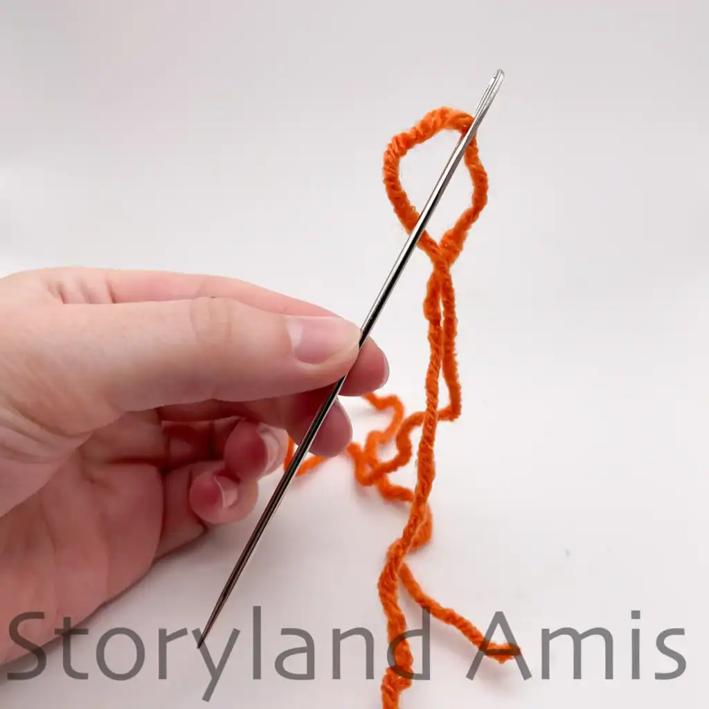 A hand holds an extra-long tapestry needle threaded with orange yarn.