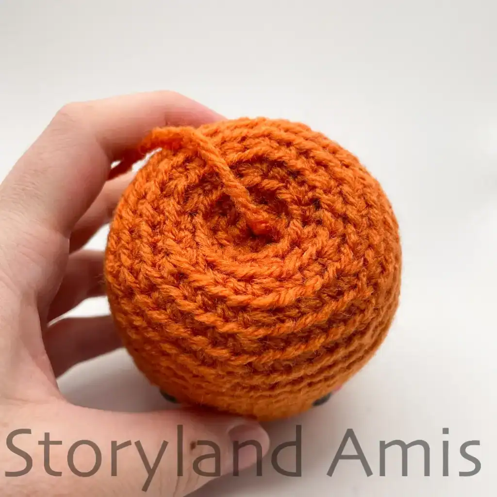 A close-up of the top of an amigurumi pumpkin. The pumpkin has a piece of yarn coming out of the top.