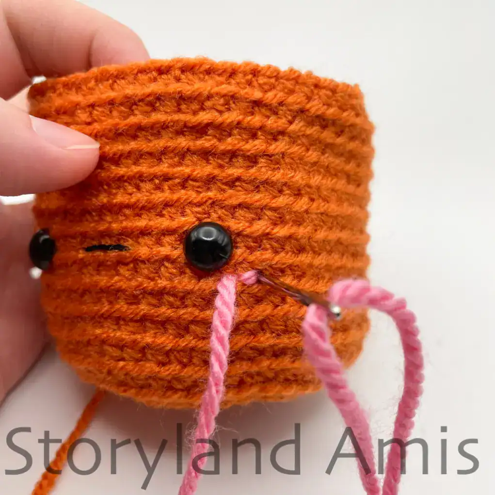 A hand demonstrates how to embroider cheeks onto the face of an amigurumi pumpkin using pink yarn.