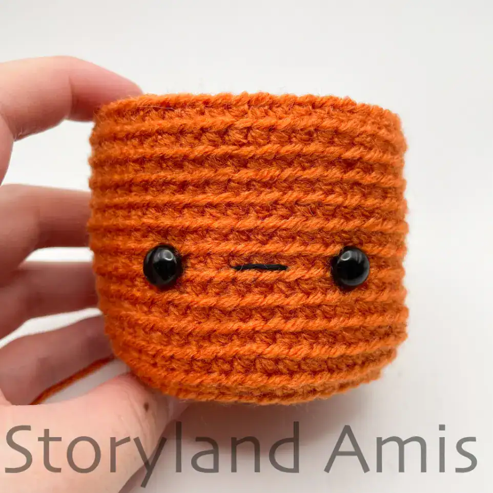 A hand holds up a partially finished crocheted pumpkin with an embroidered mouth and plastic safety eyes.